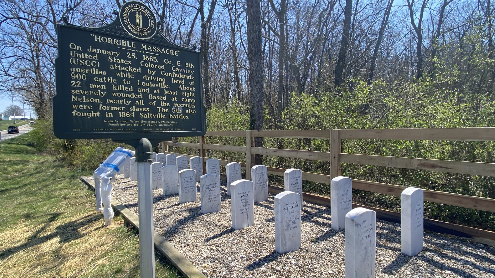 A series of white military-style headstones are placed close to one another in the ground against a fence and bushes. A state historical marker is seen in the foreground.