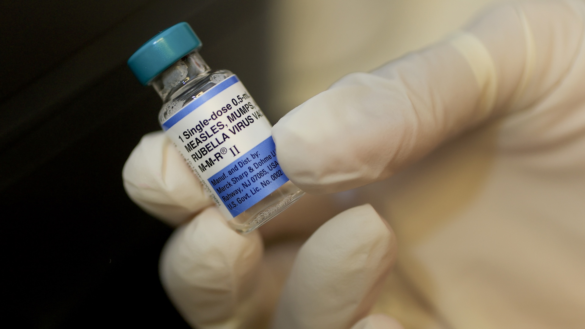 A close-up of a gloved hand holding a vial of MMR vaccine