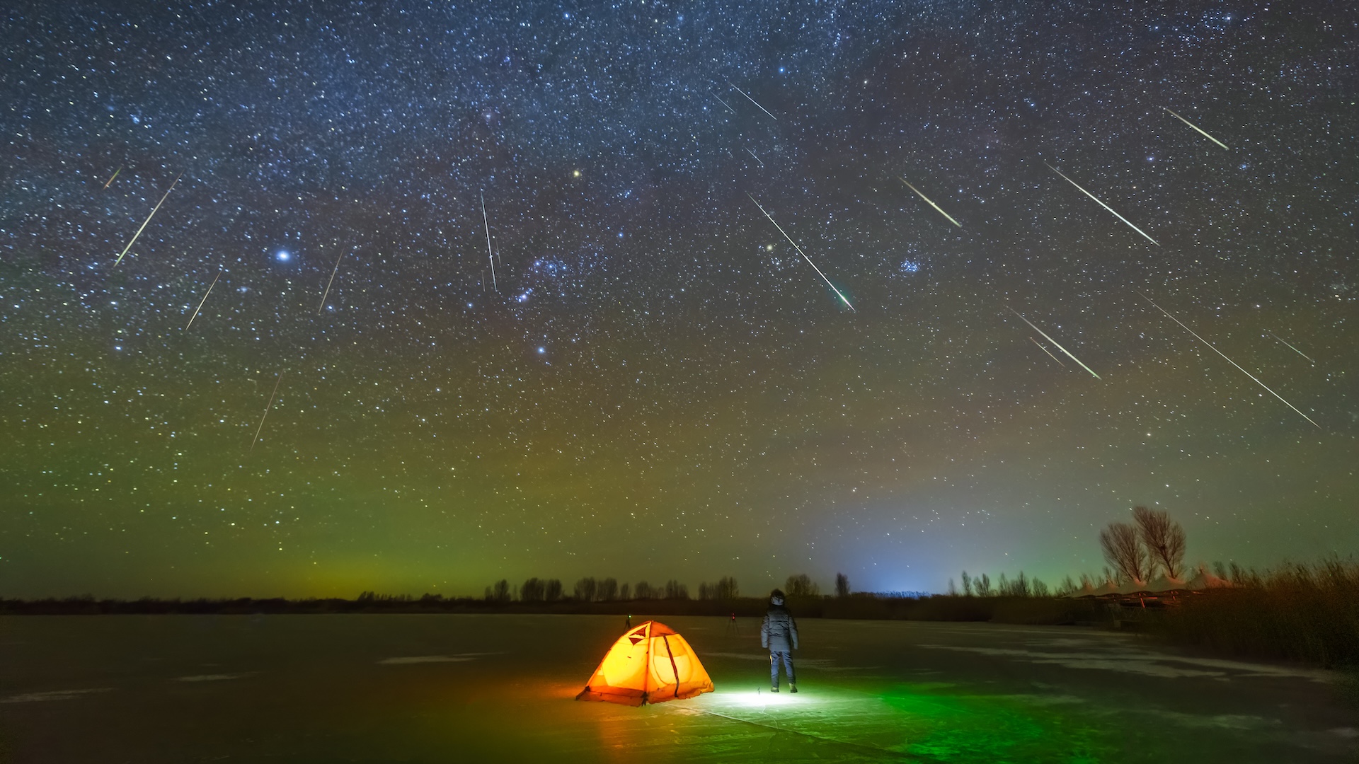 un campeur se tient devant une tente alors que le ciel nocturne est éclairé par une pluie de météores