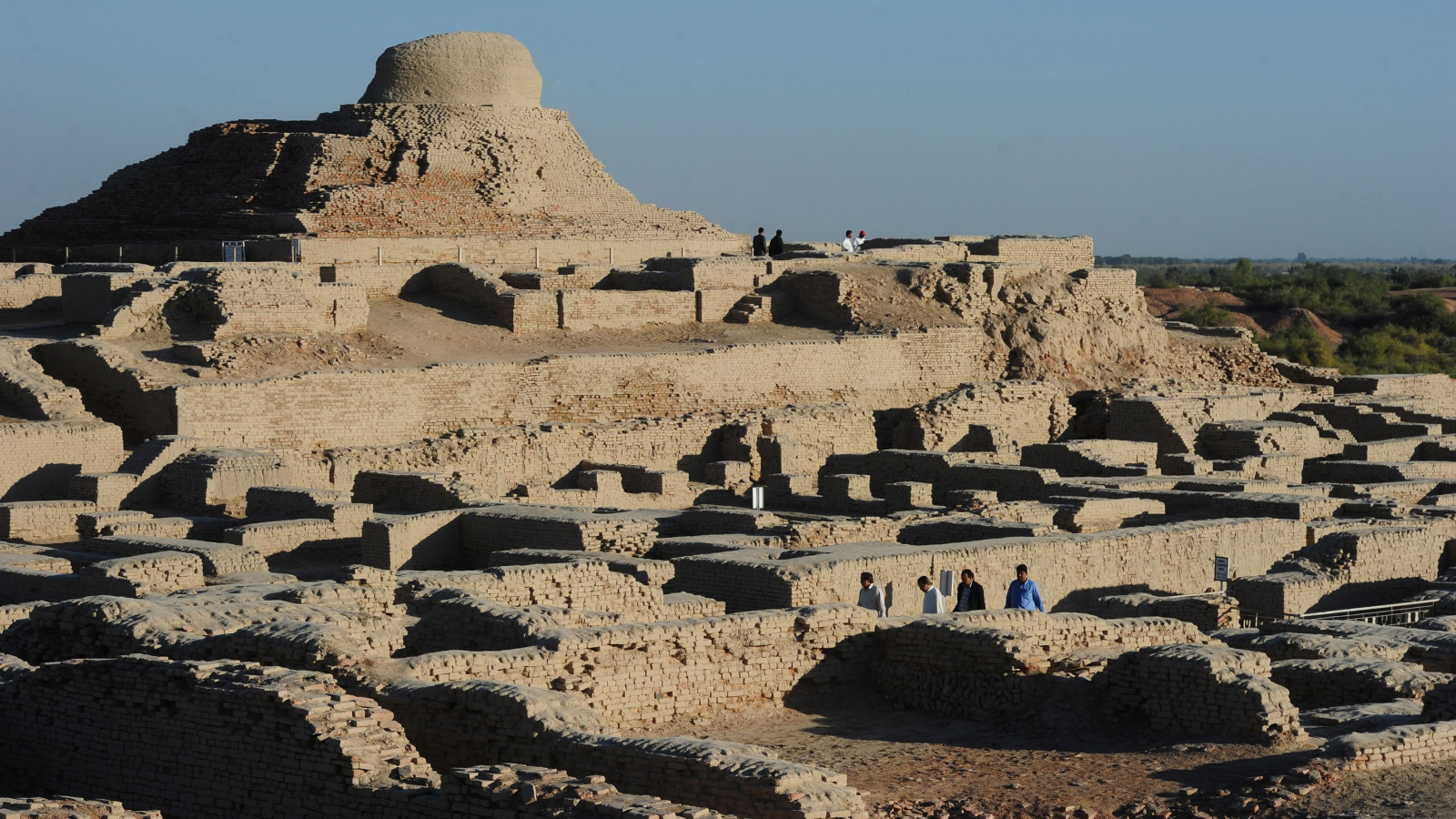 In this photograph, visitors walk through the UNESCO World Heritage archeological site of Mohenjo Daro. The image shows a complex of beige, stone structures