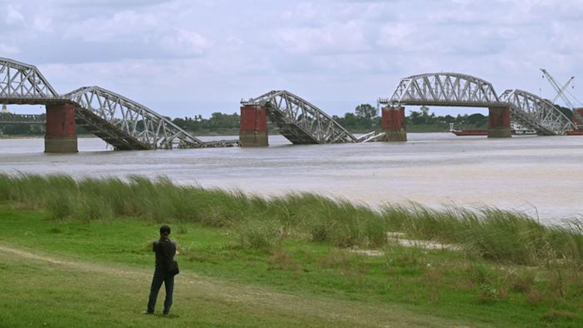 Photo taken from the grassy shore of the Irrawaddy River showing the collapsed Ava Bridge. A person stands in the foreground with their back facing the camera, with the collapsed bridge partially in the river in the distance.