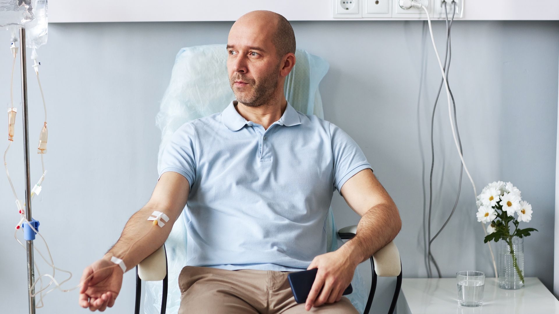 Stock photo of a middle aged white male sitting in a chair in a medical facility. He is facing the camera but looking into the distance to the right. His right arm is hooked up to an IV.