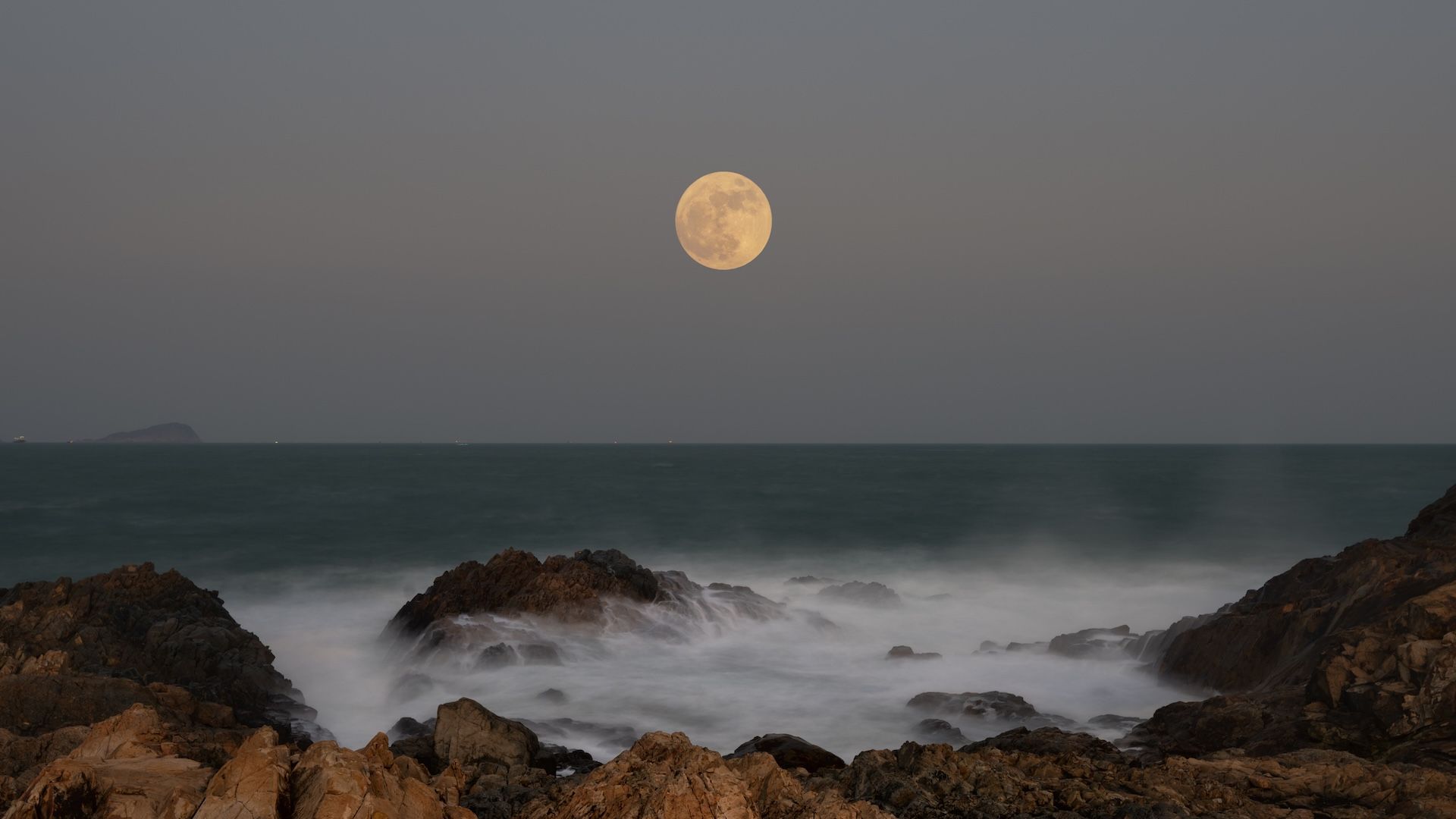 Photo of a full moon over the ocean, taken from rocky shore. Waves can be seen crashing over the rocks in the foreground/bottom of the image.