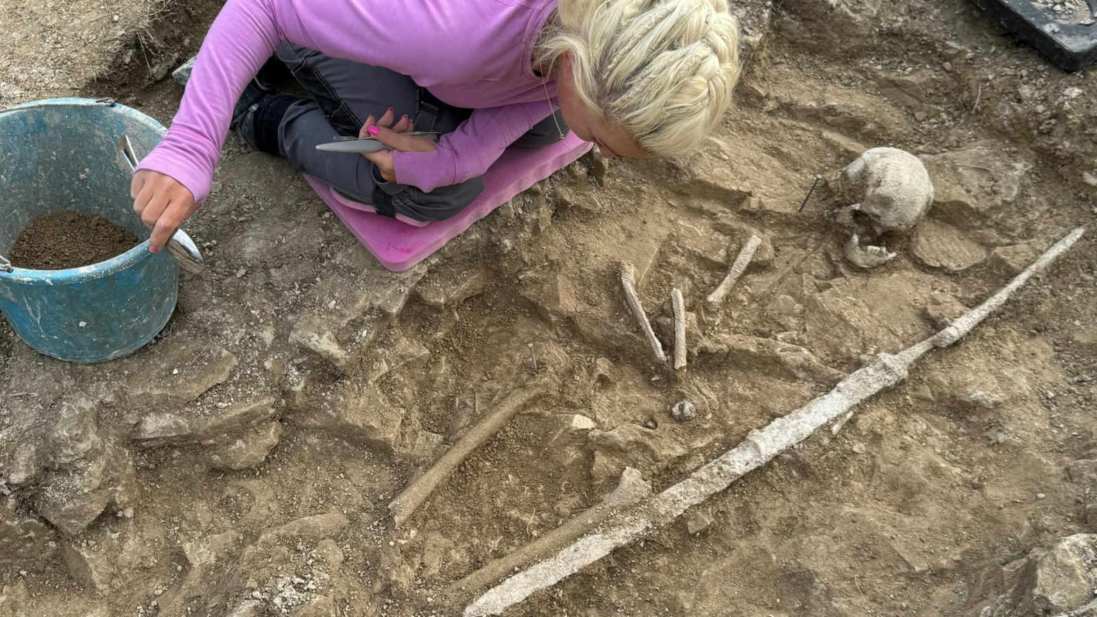A woman uncovering a dirt grave that has a skull and some long bones.