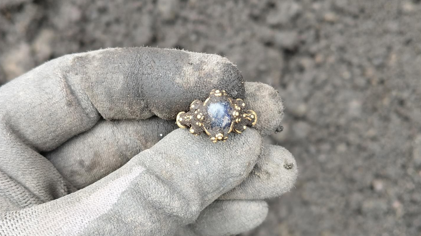 a person holds a gold ring covered in dirt with their fabric-gloved hand