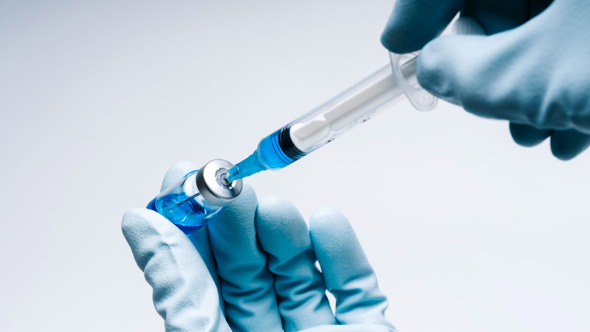 A close up shows two hands covered in blue latex gloves. The left is holding a small bottle containing blue liquid while the right hand uses a small syringe to pull out the liquid from the bottle
