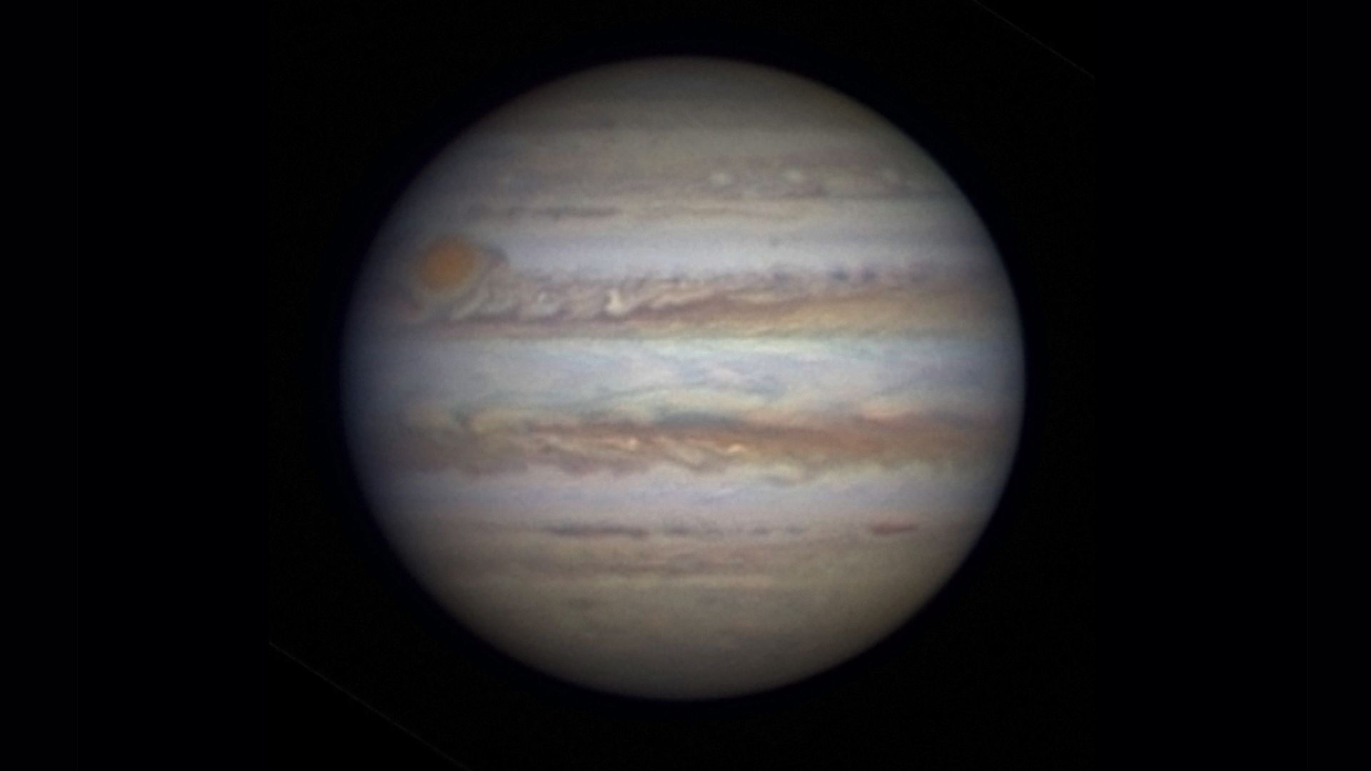 Photo de Jupiter vue à travers un télescope. Une grande sphère blanche entourée d’anneaux tourbillonnants de rouge et de brun sur un fond noir. 