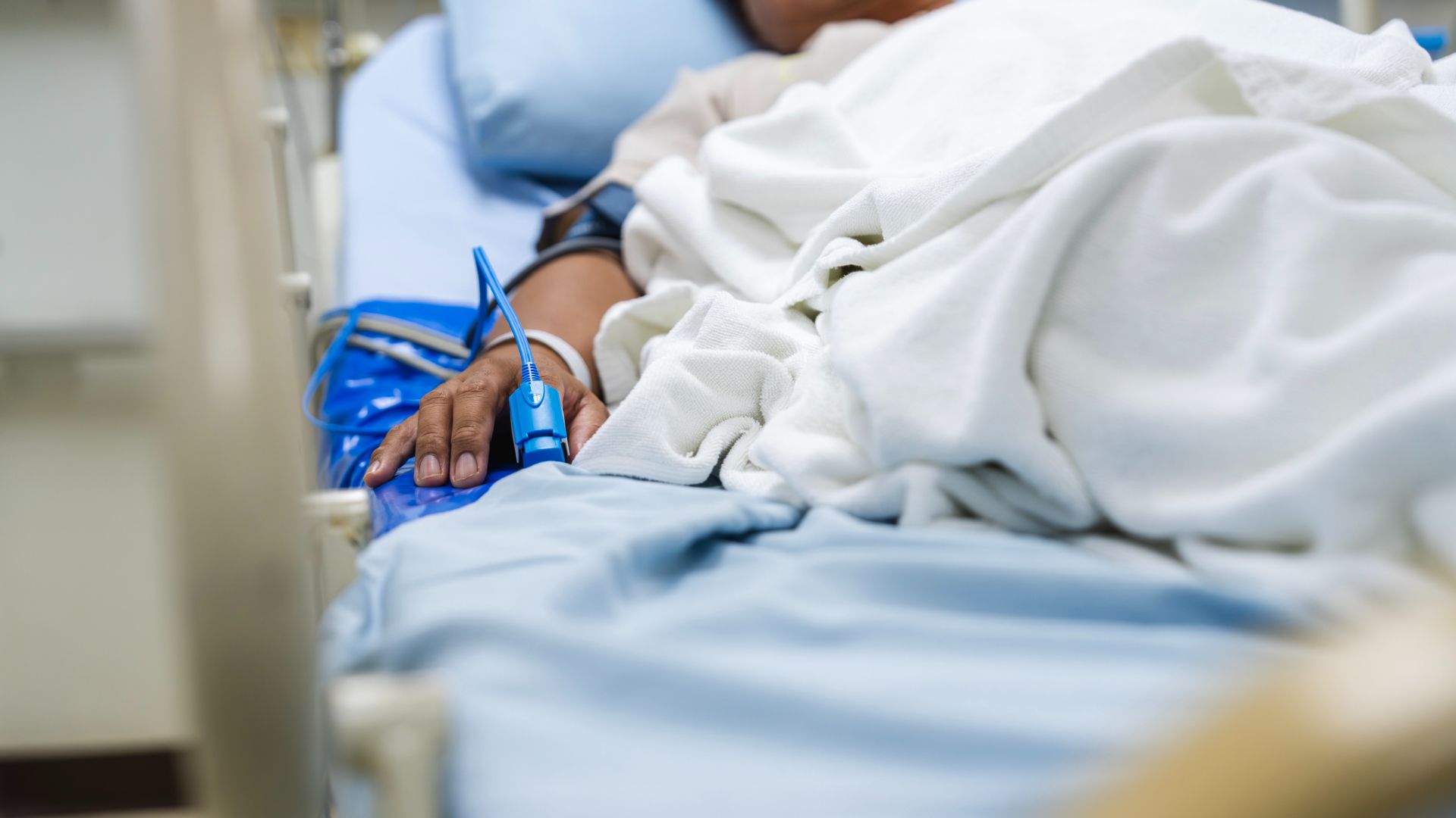 A close up of the right side of a tan person lying in a moving hospital bed, with a blue blood monitor clamped on their right index finger. Their body is covered by white sheets on a blue mattress.
