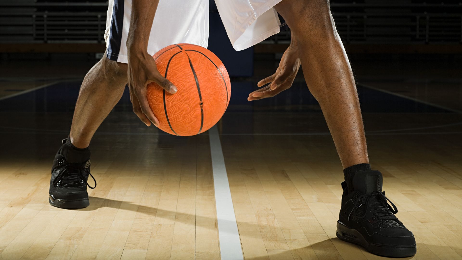 A close up of a man's lower legs, with both feet wearing tall black sneakers and black socks. He wears white basketball shorts and is bouncing a basketball between his hands while standing on a midline on a wooden basketball court