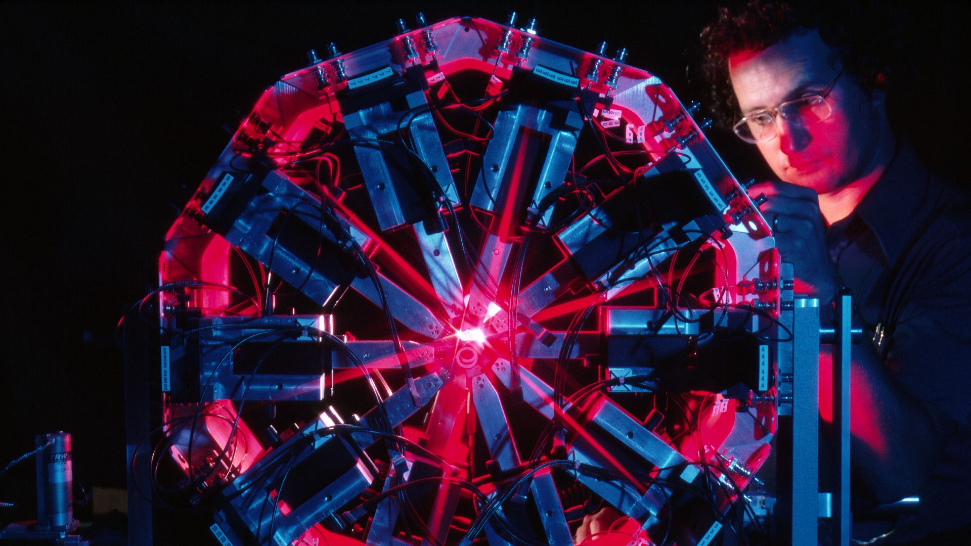 A man with glasses and a blue shirt stands behind a Ferris-wheel looking contraption with red and blue lighting in a dark room