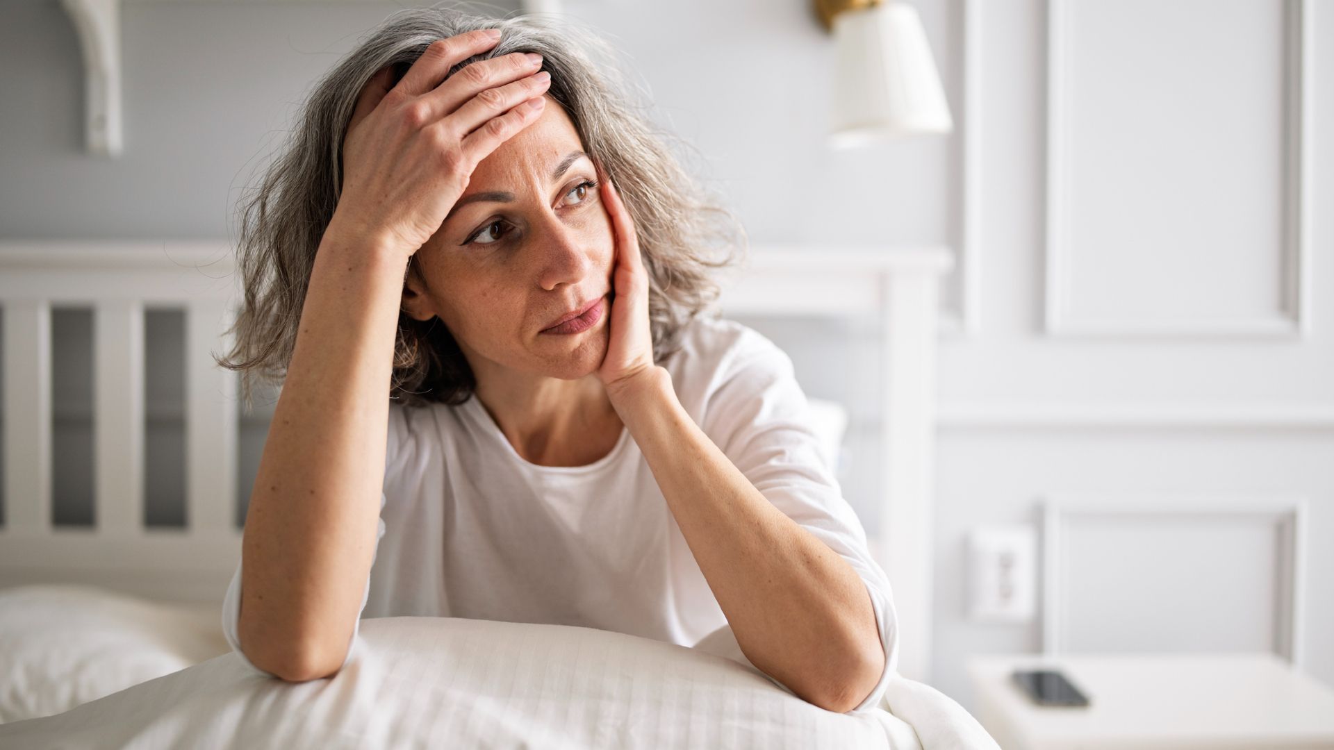 A woman with short gray wavy hair wearing a white t-shirt holds her forehead in one hand, her elbows propped up on a pillow on her bed.