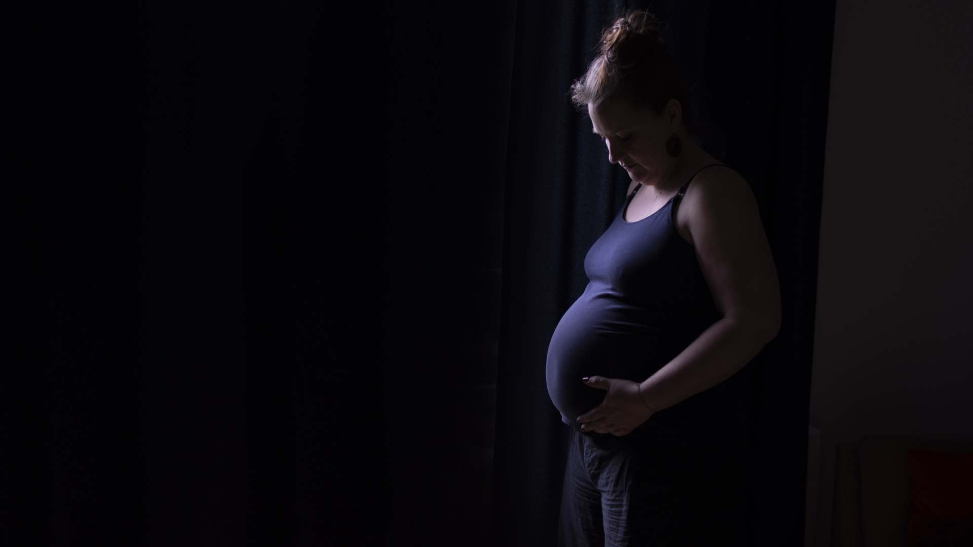 Une femme enceinte vêtue d'un débardeur violet regarde son ventre debout devant un fond noir
