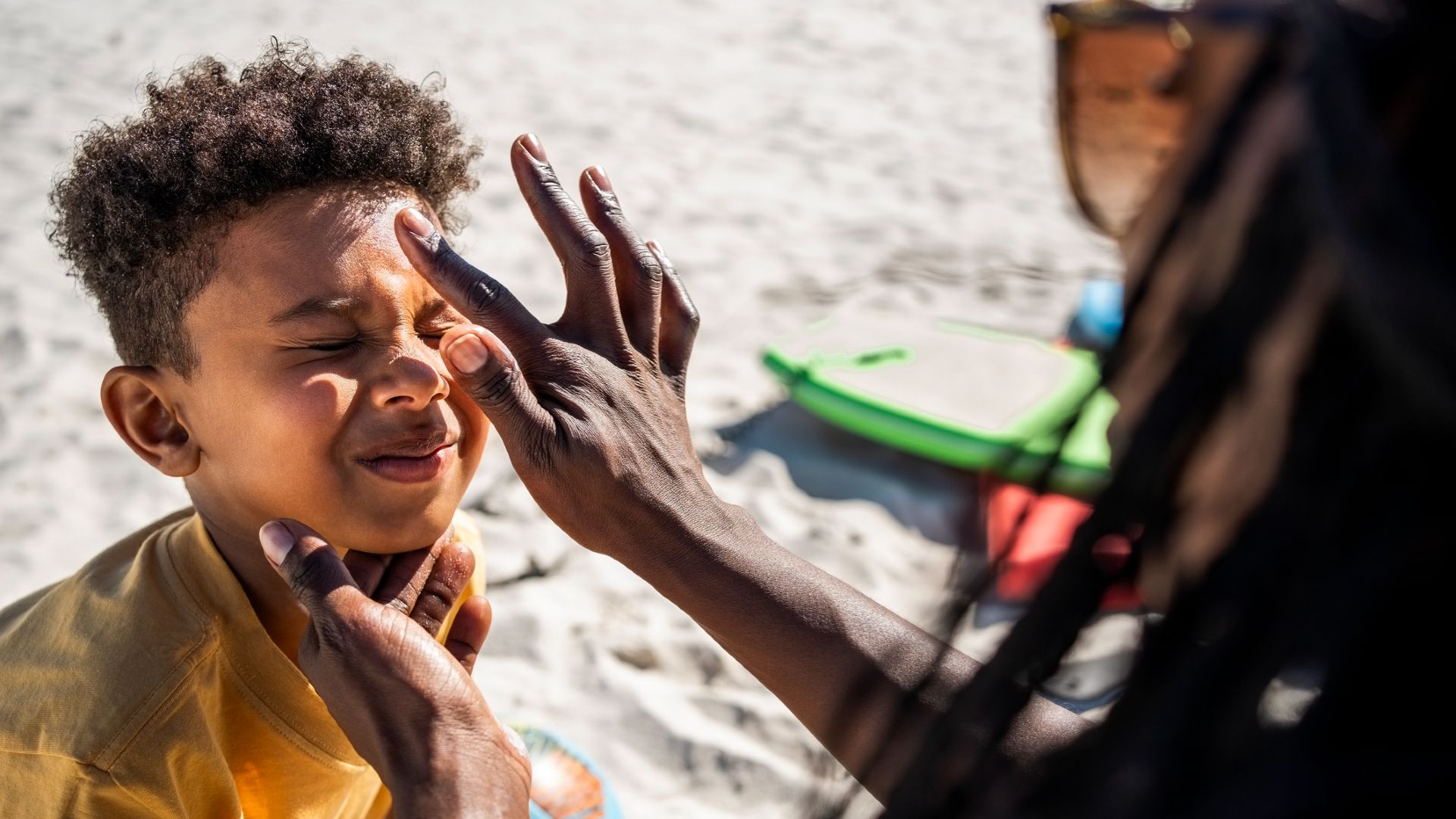 A woman wearing sunglasses with long hair applies a dot of sunscreen to a young boy who scrunches up his face in response