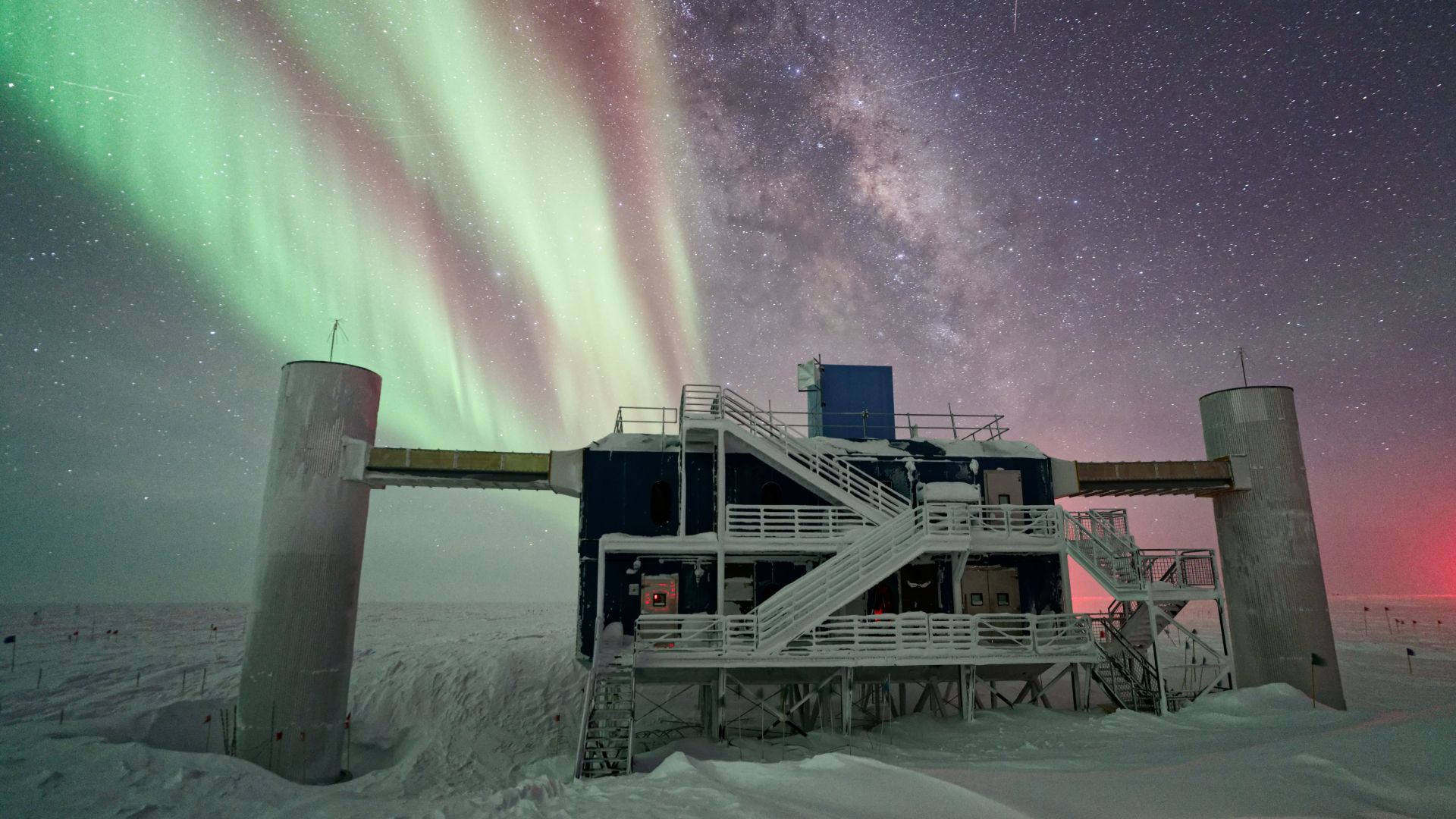 A concrete structure with large pillars and a metal staircase looms over a snowy landscape with green and red auroras illuminating the night sky