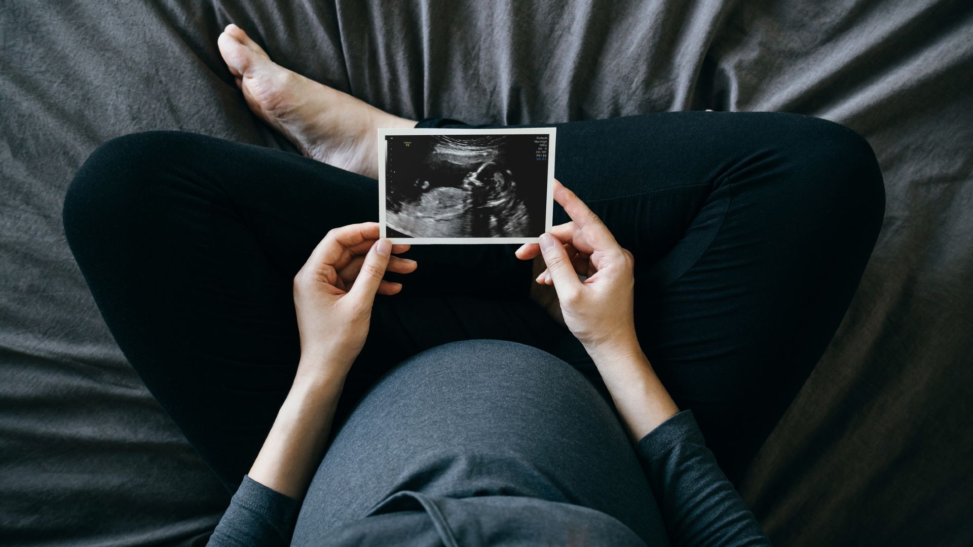 A top down shot of a pregnant person wearing a blue shirt and black pants sitting cross-legged on a blue bed, holding an ultrasound image in front of them.