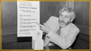 A black and white photo shows a man with long hair and a beard wearing a suit and patterned tie pointing at a poster labeled "AIDS is everyone's problem"