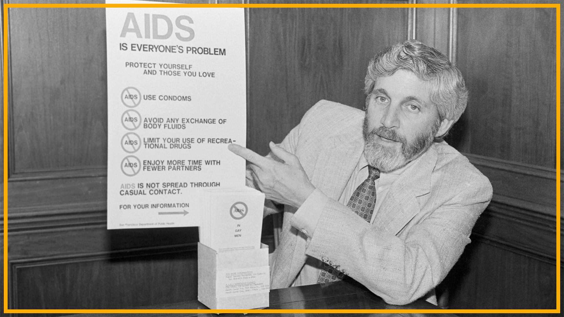 A black and white photo shows a man with long hair and a beard wearing a suit and patterned tie pointing at a poster labeled "AIDS is everyone's problem"