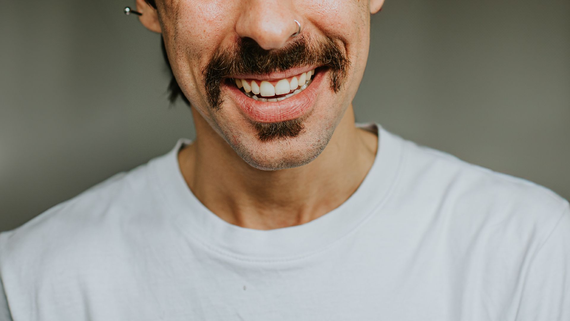 An image of the bottom half of a man's face, showing a dark moustache over a white-toothed smile. The man has an earring bar in his left ear and wears a white shirt