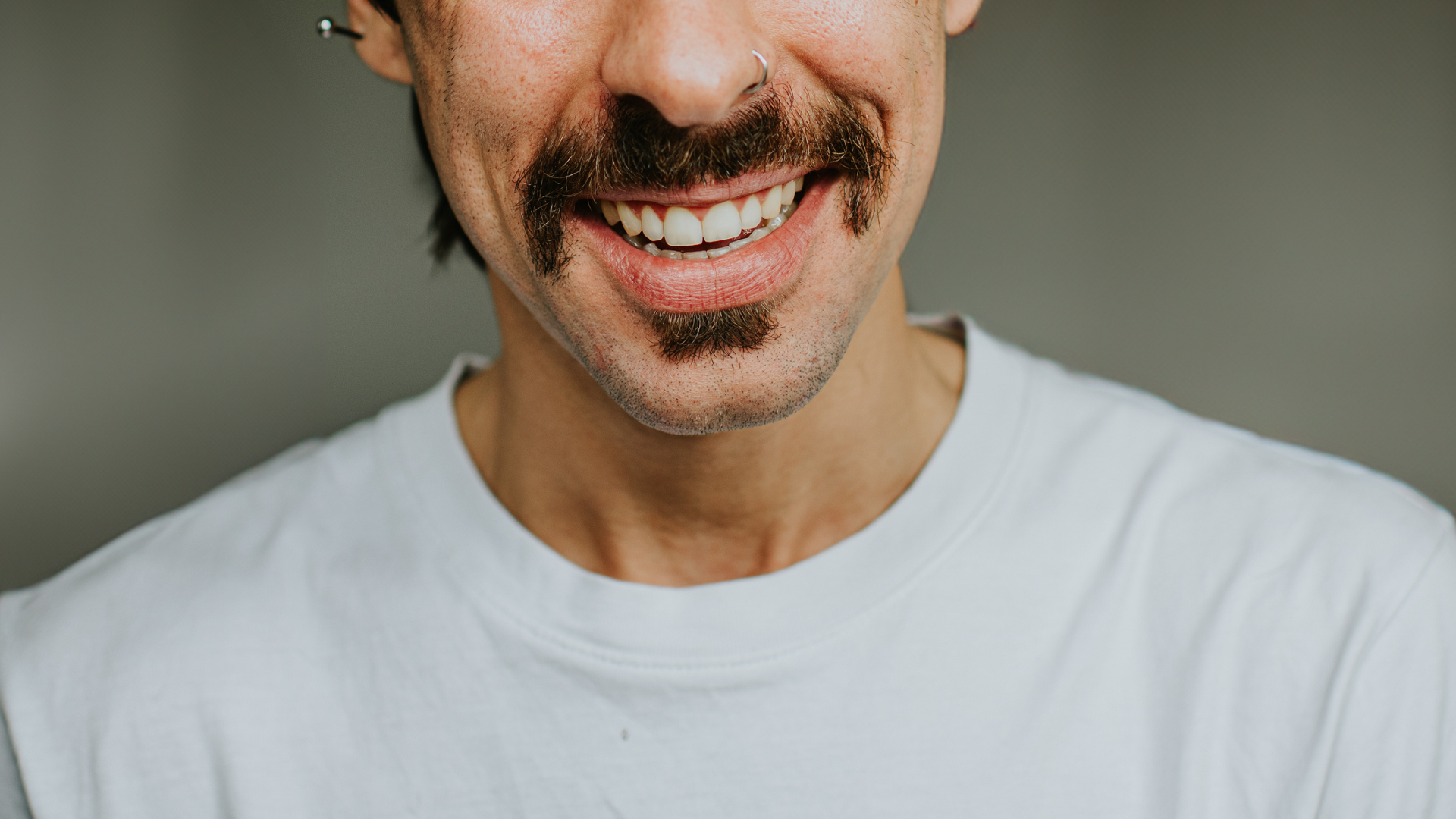 Image de la moitié inférieure du visage d'un homme, montrant une moustache sombre sur un sourire aux dents blanches. L'homme a une boucle d'oreille à l'oreille gauche et porte une chemise blanche