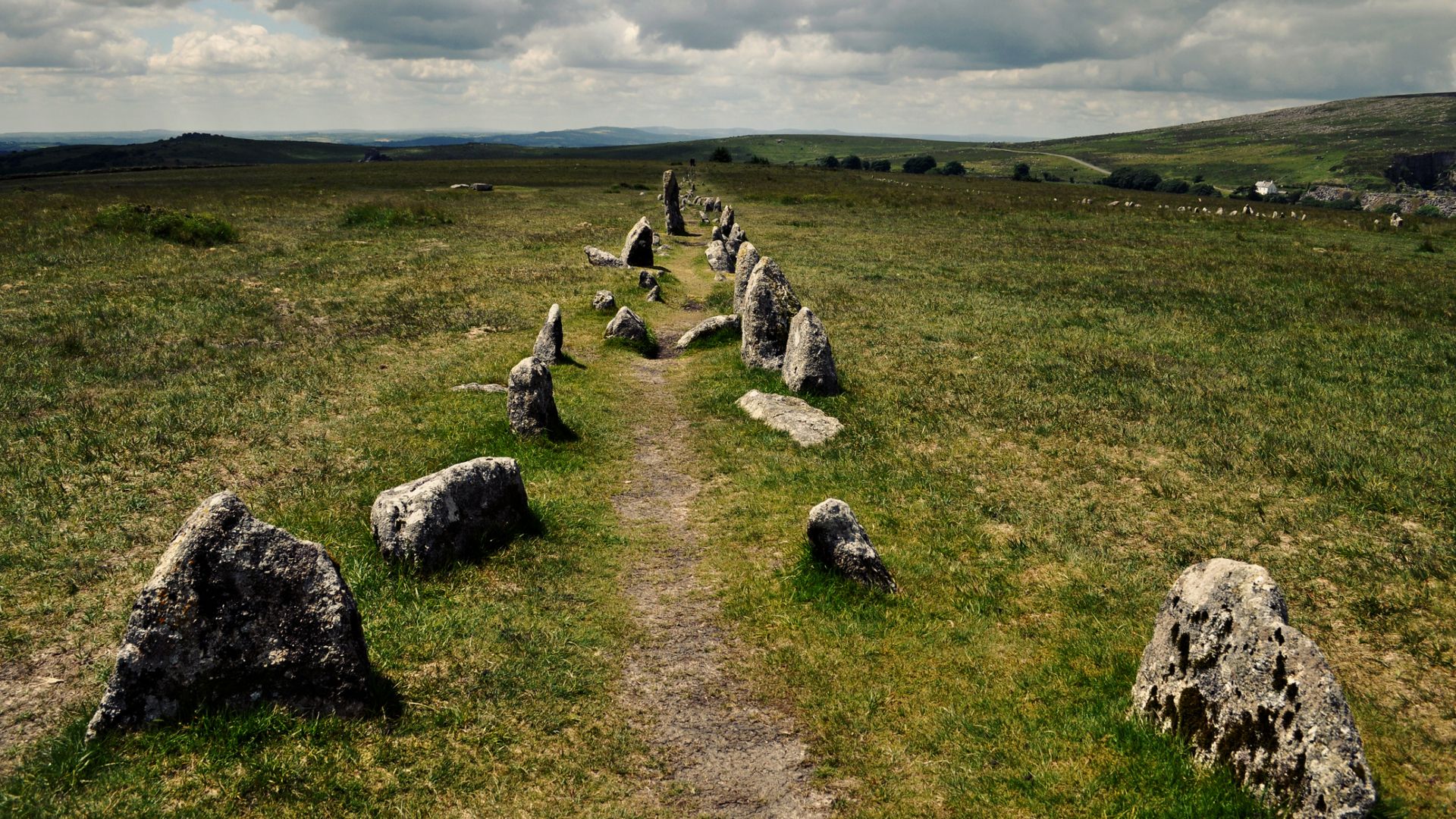 A view of lush rolling hills under a cloudy gray sky. In the foreground is a faded narrow dirt road with stones on either side of it