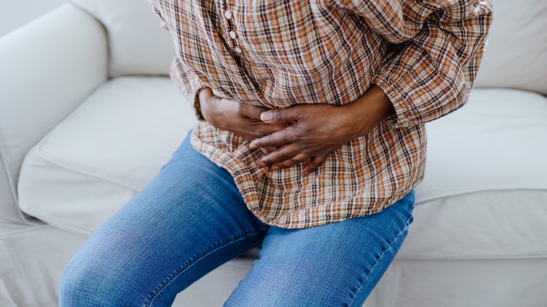 Close-up of a woman holding her hands over her pelvis in pain. She is wearing a brown checked shirt and blue denim jeans and is sat on a white couch.