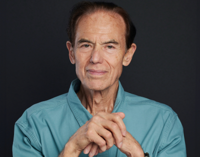 Un homme aux cheveux noirs portant une chemise boutonnée bleue regarde la caméra
