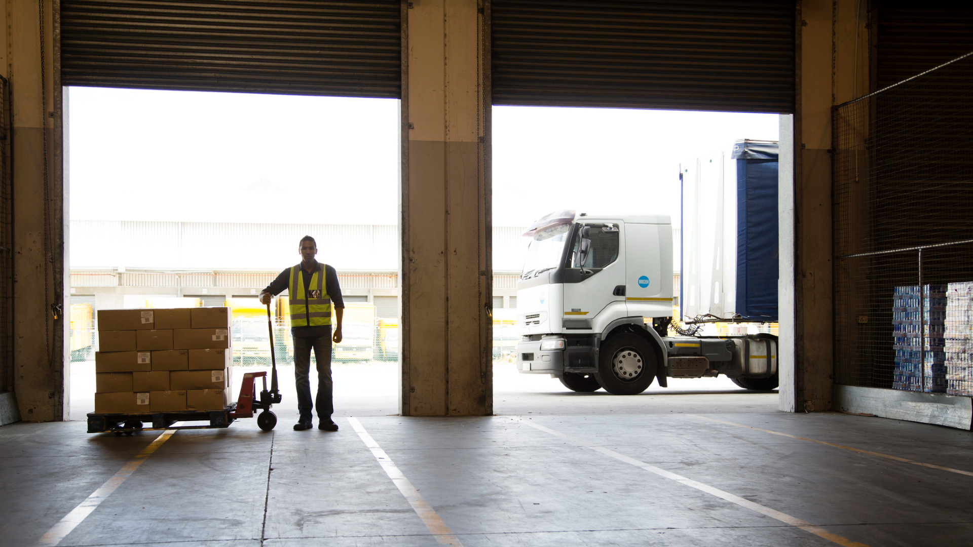 Un travailleur portant un gilet jaune regarde un entrepôt vide avec un chariot rempli de boîtes de nourriture à côté de lui et un camion en arrière-plan
