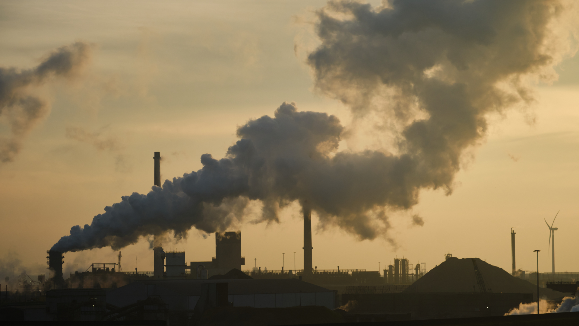Une cheminée dégage une épaisse fumée grise sur un ciel jaunâtre et smogeux, avec les bâtiments en contrebas assombris dans la brume