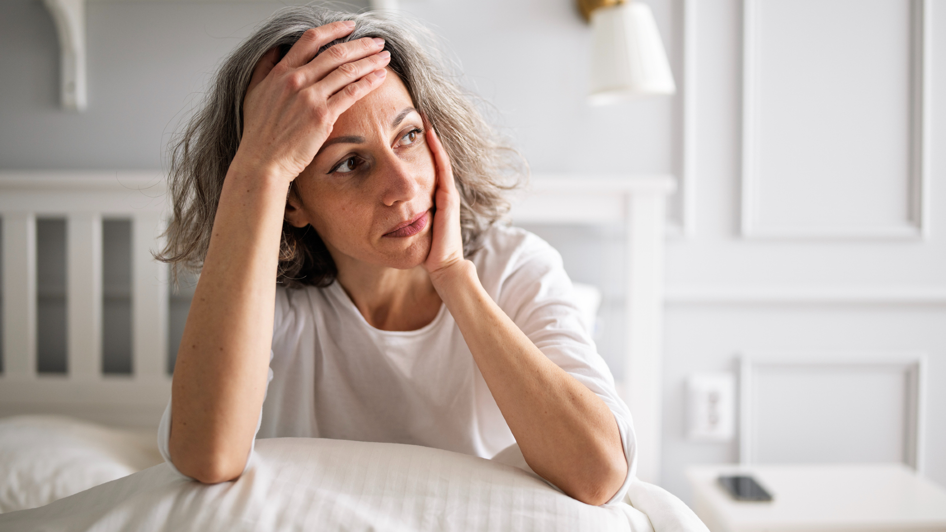 Une femme aux cheveux gris courts et ondulés portant un t-shirt blanc tient son front dans une main, ses coudes appuyés sur un oreiller sur son lit. 