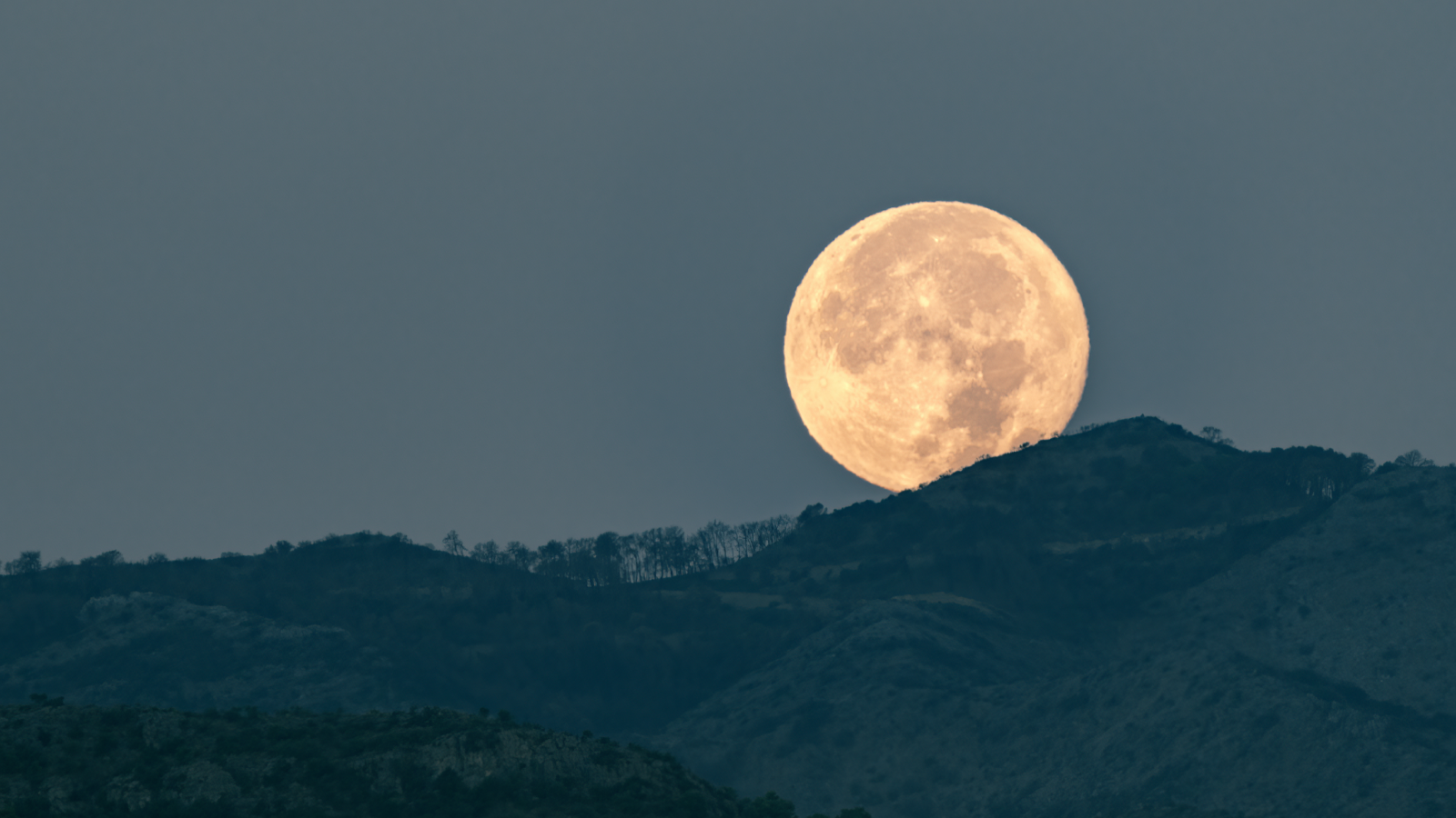 Cette photographie fige le moment exact de l'aube lorsque la pleine lune disparaît derrière les montagnes, projetant une lumière sereine et éthérée sur le paysage. Le spectacle tranquille de la nature se déroule alors que la lune fait ses adieux silencieux sur fond de monde éveillé.