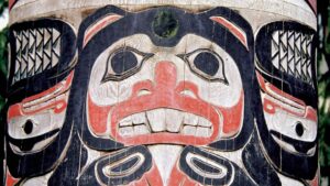 A large black, red and white wood carved totem pole shows a large face with black eyes and white teeth with feathers on either side.