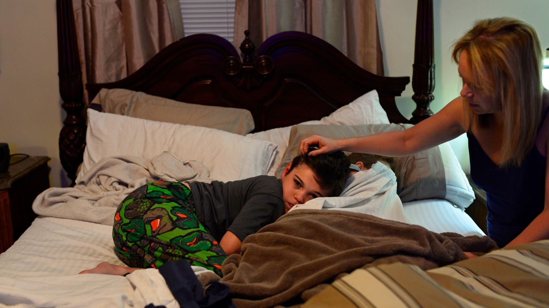 A child wearing a gray shirt and patterned green pants curls up on a bed with his mother, a blond woman with long straight hair, caressing his head