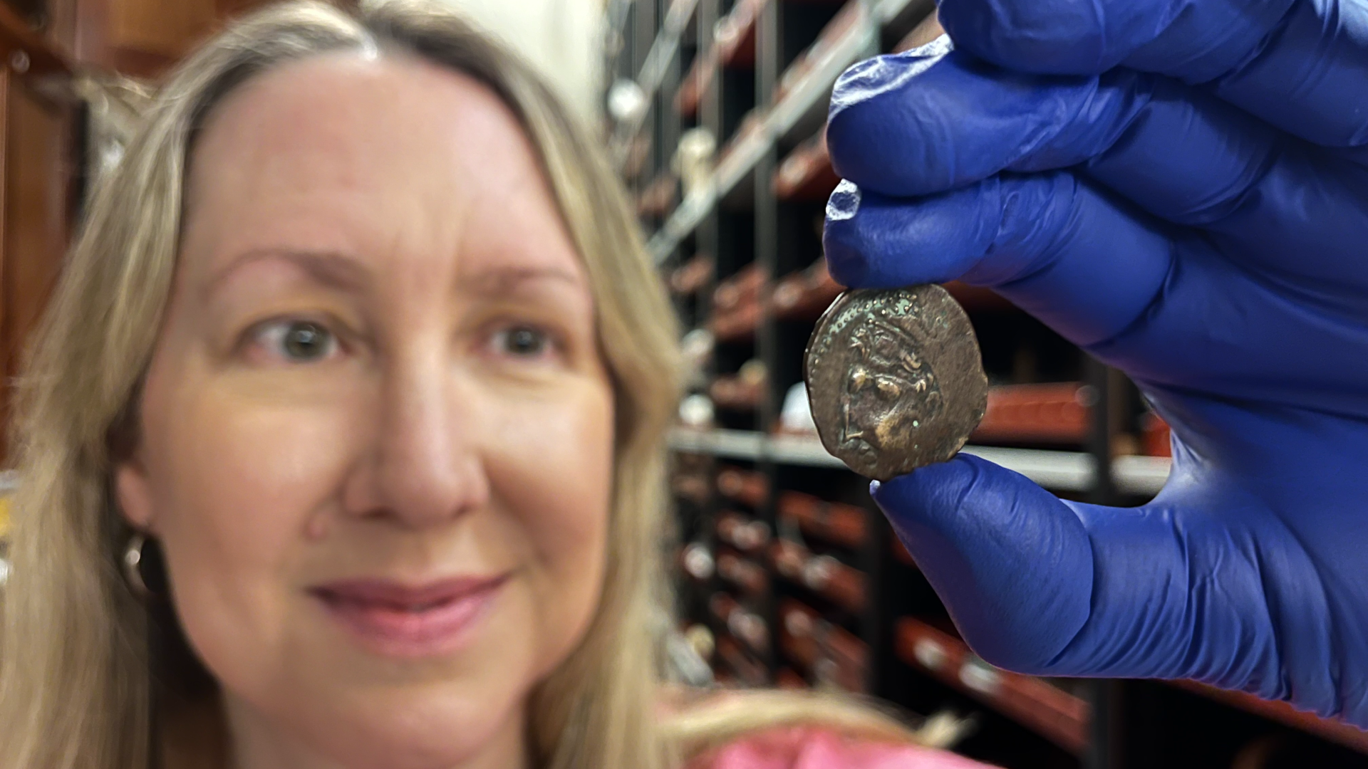 A blond woman wearing blue disposable gloves shows off the front of the ancient coin.