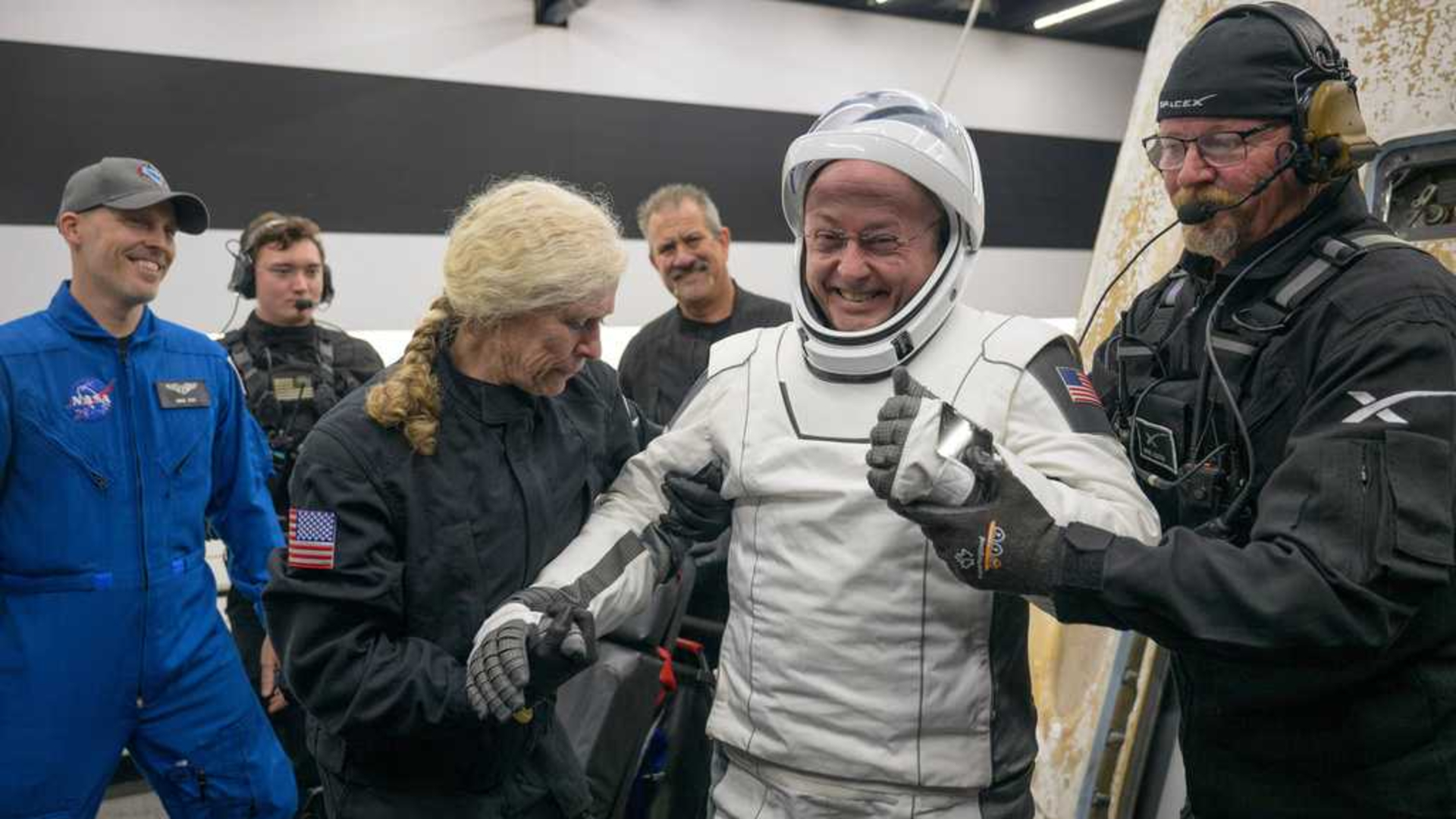 Un homme vêtu d’une combinaison spatiale blanche est accompagné de deux personnes portant une combinaison noire.