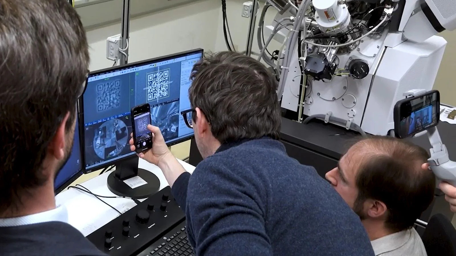 Trois hommes se blottissent autour d’un écran d’ordinateur dans un laboratoire à côté d’une grande machine. L'homme au milieu, vêtu d'une manche longue bleu marine, tient un téléphone devant le moniteur et scanne un code QR. 