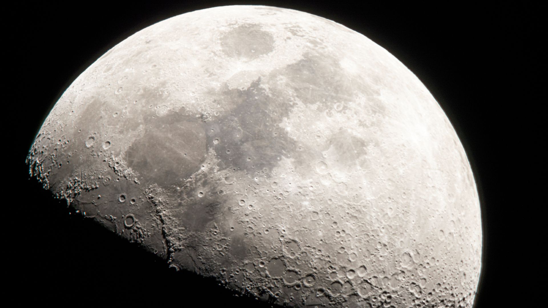 A close up of the moon in the darkness of space, its craters and dark maria visible against the gray surface, while its bottom half is covered in shadow.
