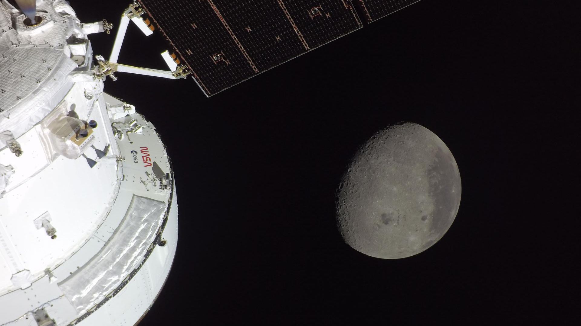 A gray sphere in the darkness of space is seen from a white spacecraft with solar panels to the left of the image.
