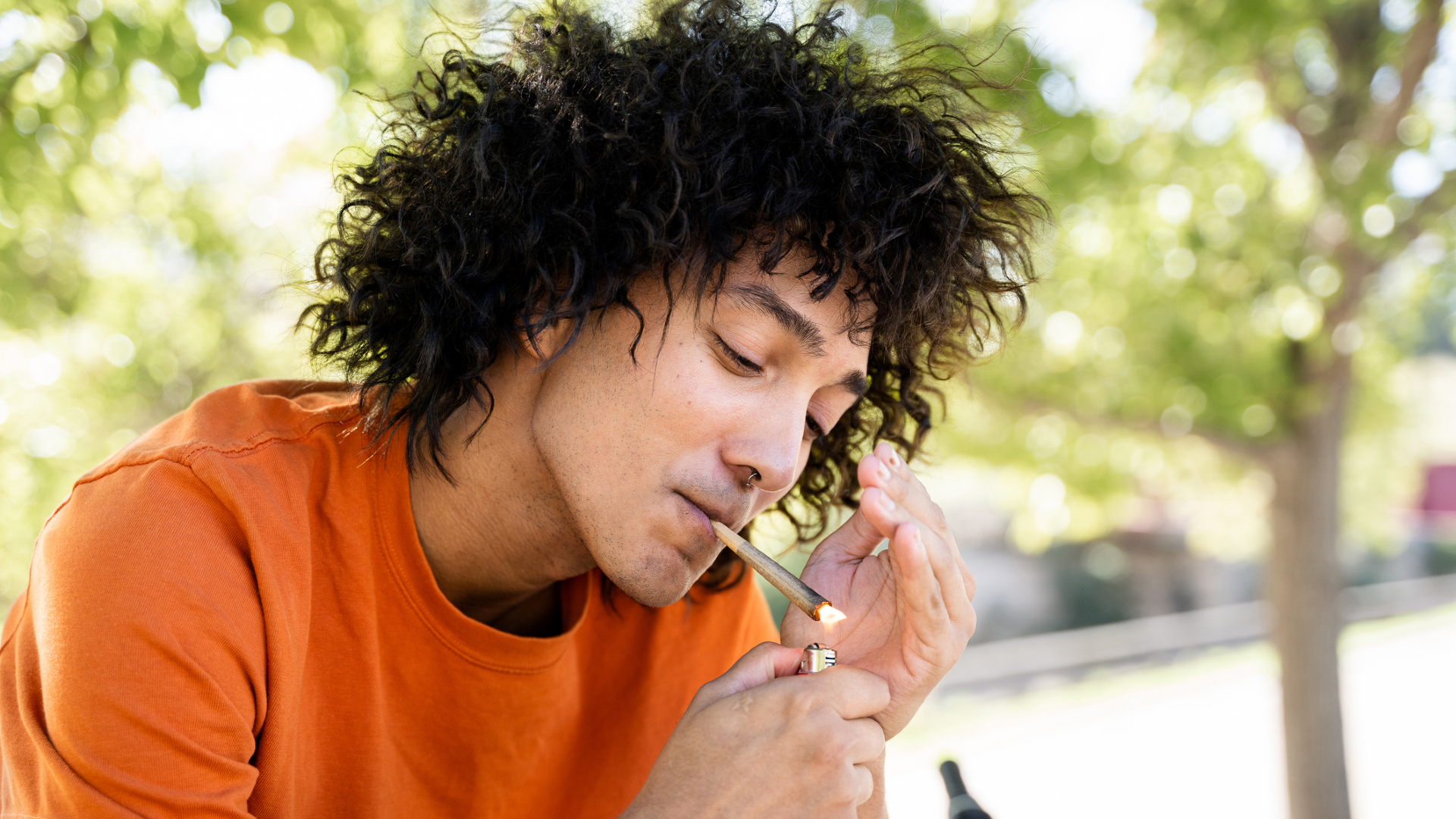 Un homme aux longs cheveux noirs ondulés portant une chemise orange allume un joint avec un briquet