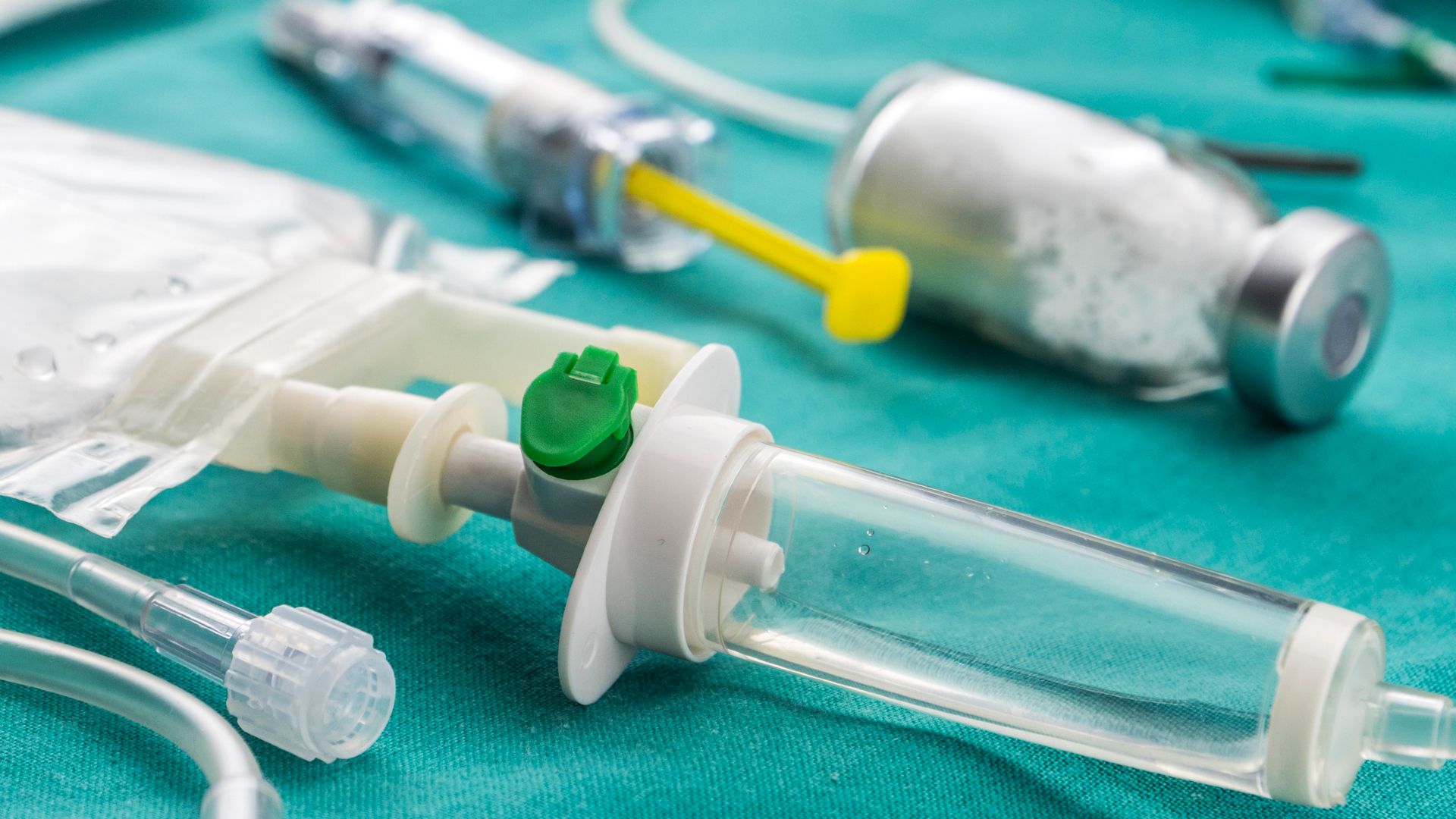 A close up of a series of medicine bottles, IV drips and syringes sitting on a turquoise sheet.