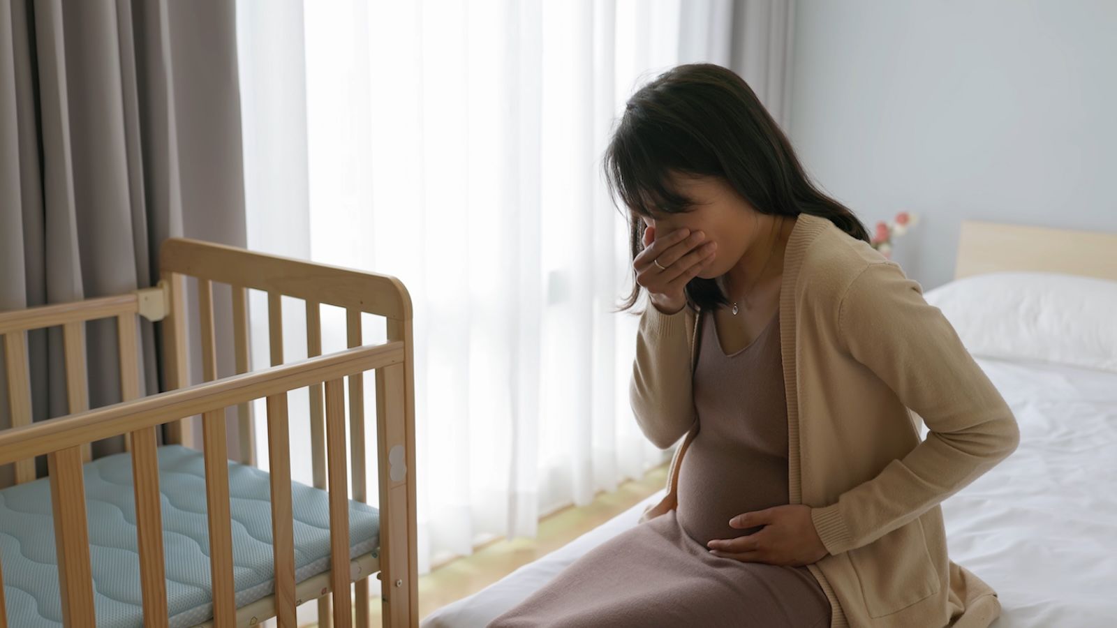 Pregnant person on bed next to crib holding mouth and stomach