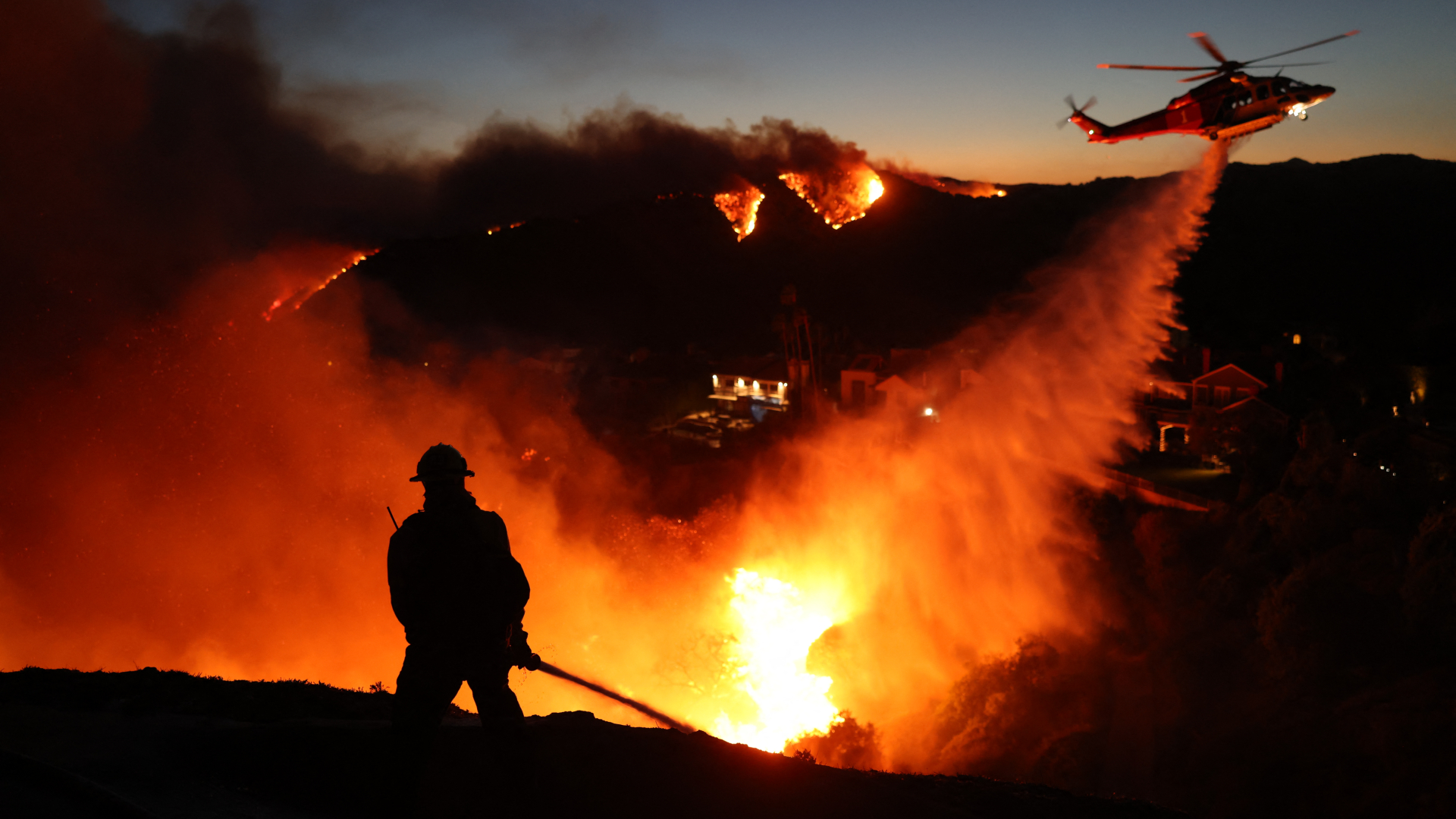 Un pompier se profile dans l'incendie orange et jaune brillant d'un incendie de forêt alors qu'un hélicoptère au-dessus déverse un jet d'eau en dessous.