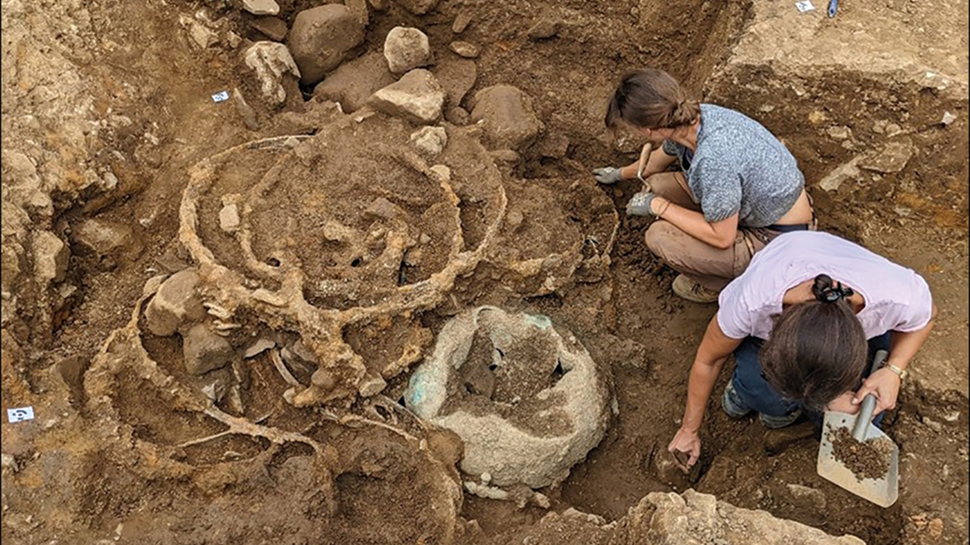 Vue d'un site de fouilles, où deux femmes accroupies à droite recueillent des échantillons de pierres à moitié déterrées.