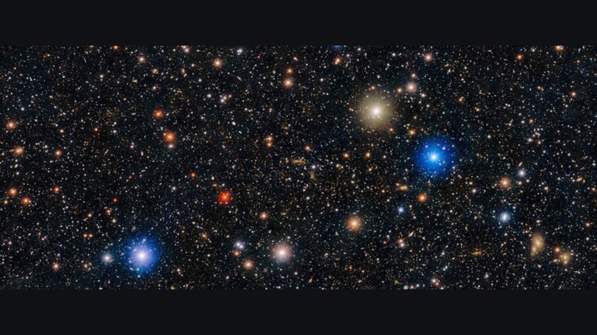 Image de l'espace montrant un ciel noir avec des centaines de points blancs et quelques points rouges, orange, dorés et bleus entourés de halos de lumière. 