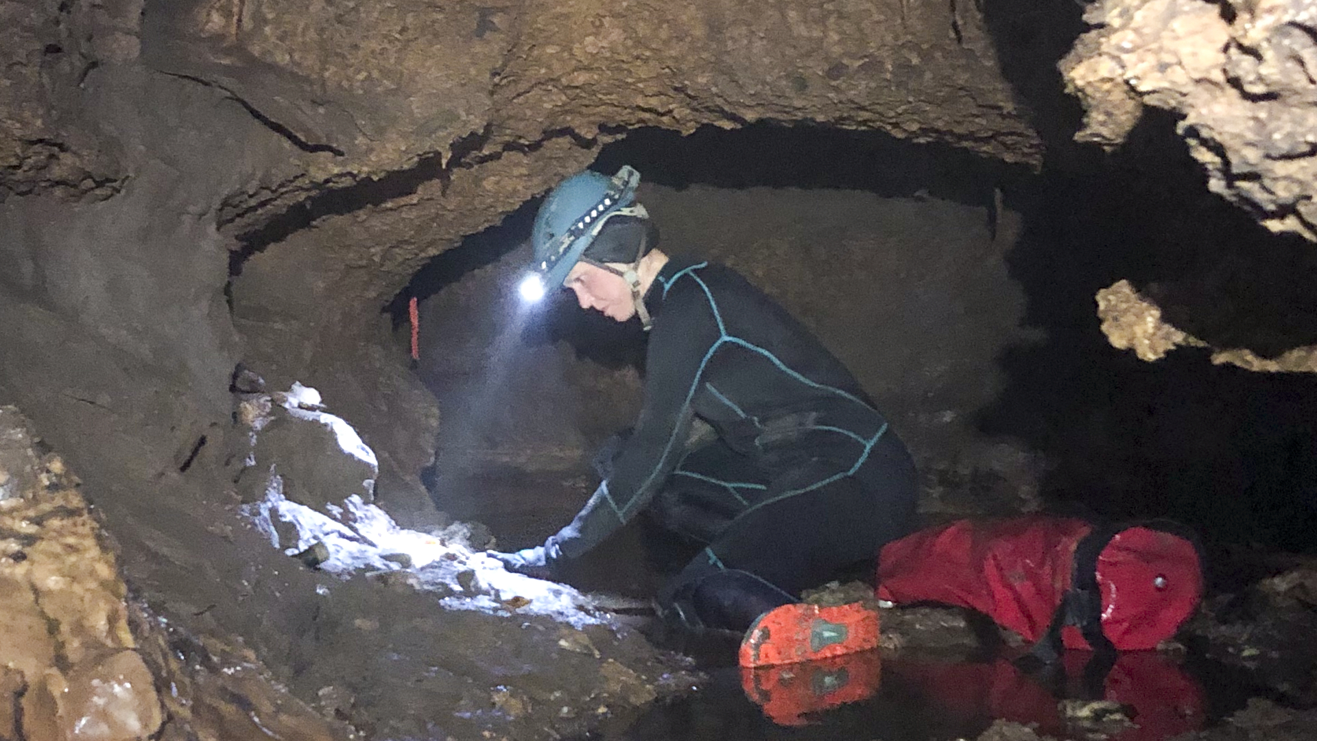 Une personne vêtue d’un costume noir et d’un casque de sécurité avec une lampe de poche se penche sur le mur circulaire d’une petite grotte. 