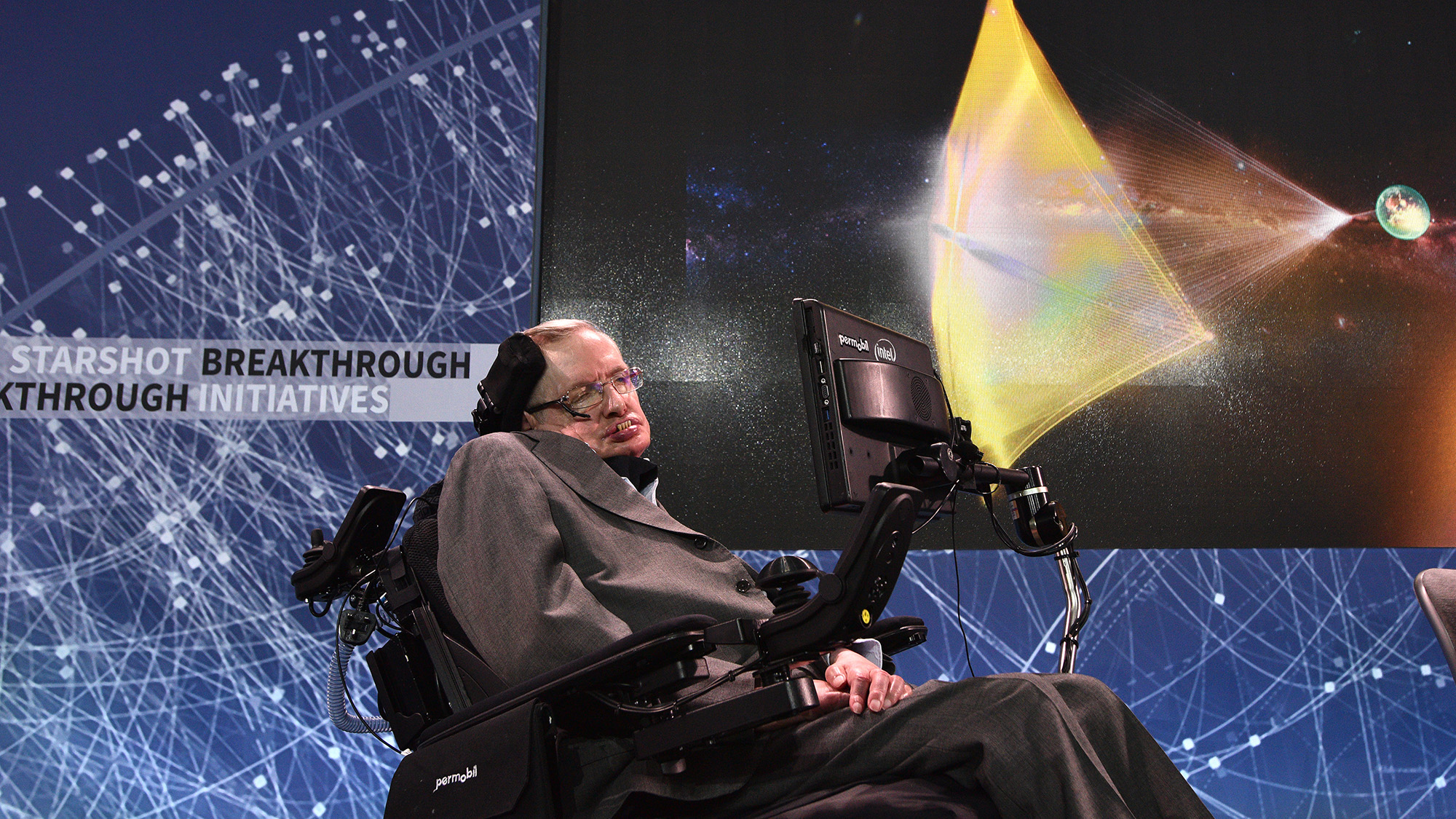 Un homme dans un fauteuil roulant électronique se tient devant un écran de projection sur lequel figurent diverses images spatiales.