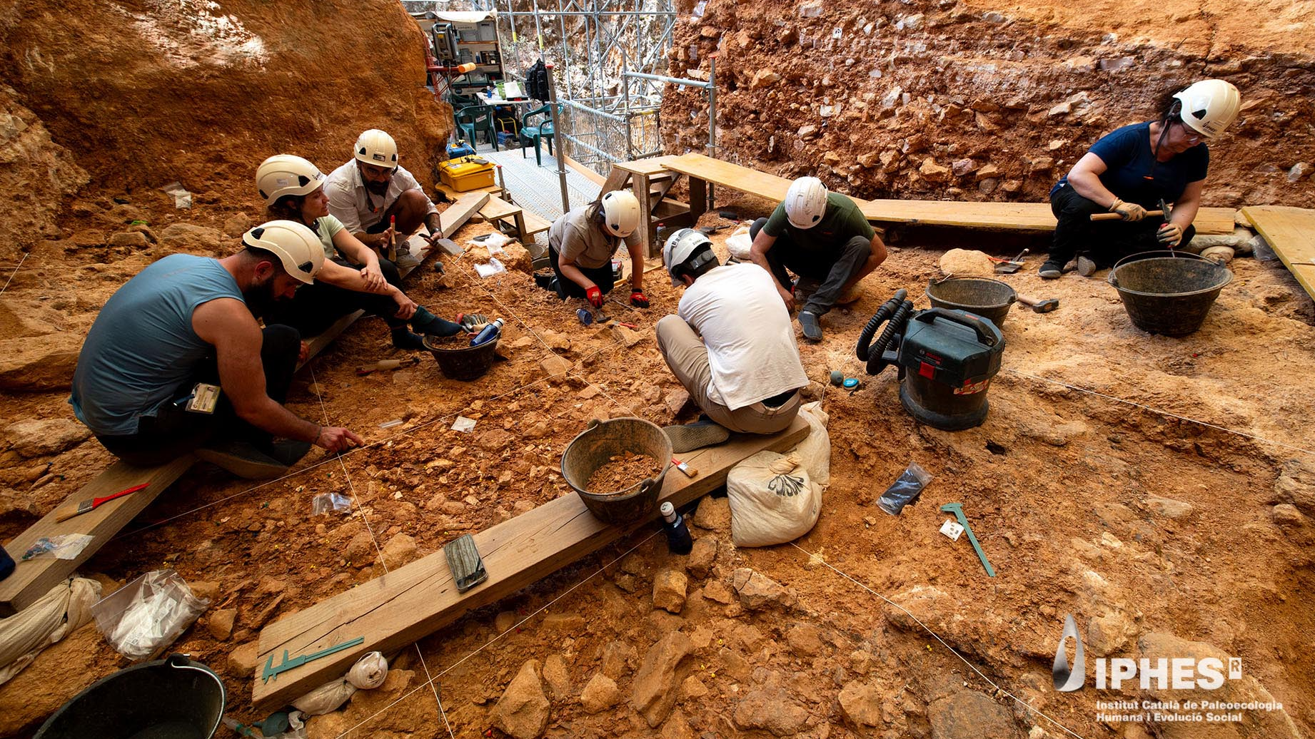 seven adults with white hard hats excavate in orange-colored soil in a cave