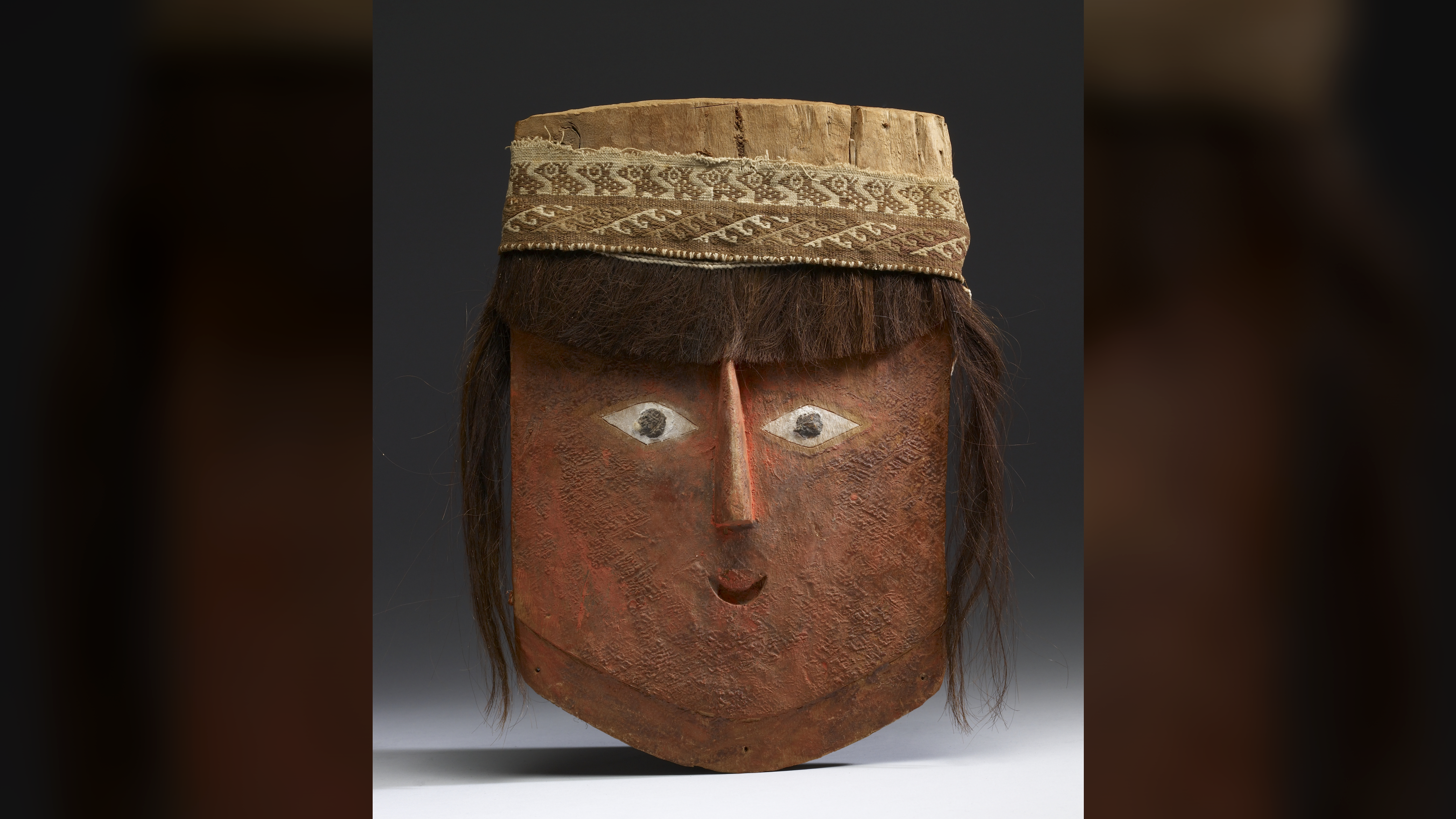 un masque en bois peint en rouge en forme de visage humain stylisé avec des cheveux humains attachés