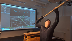 a man with light skin wears a motion-capture suit and swings a large stick over his head