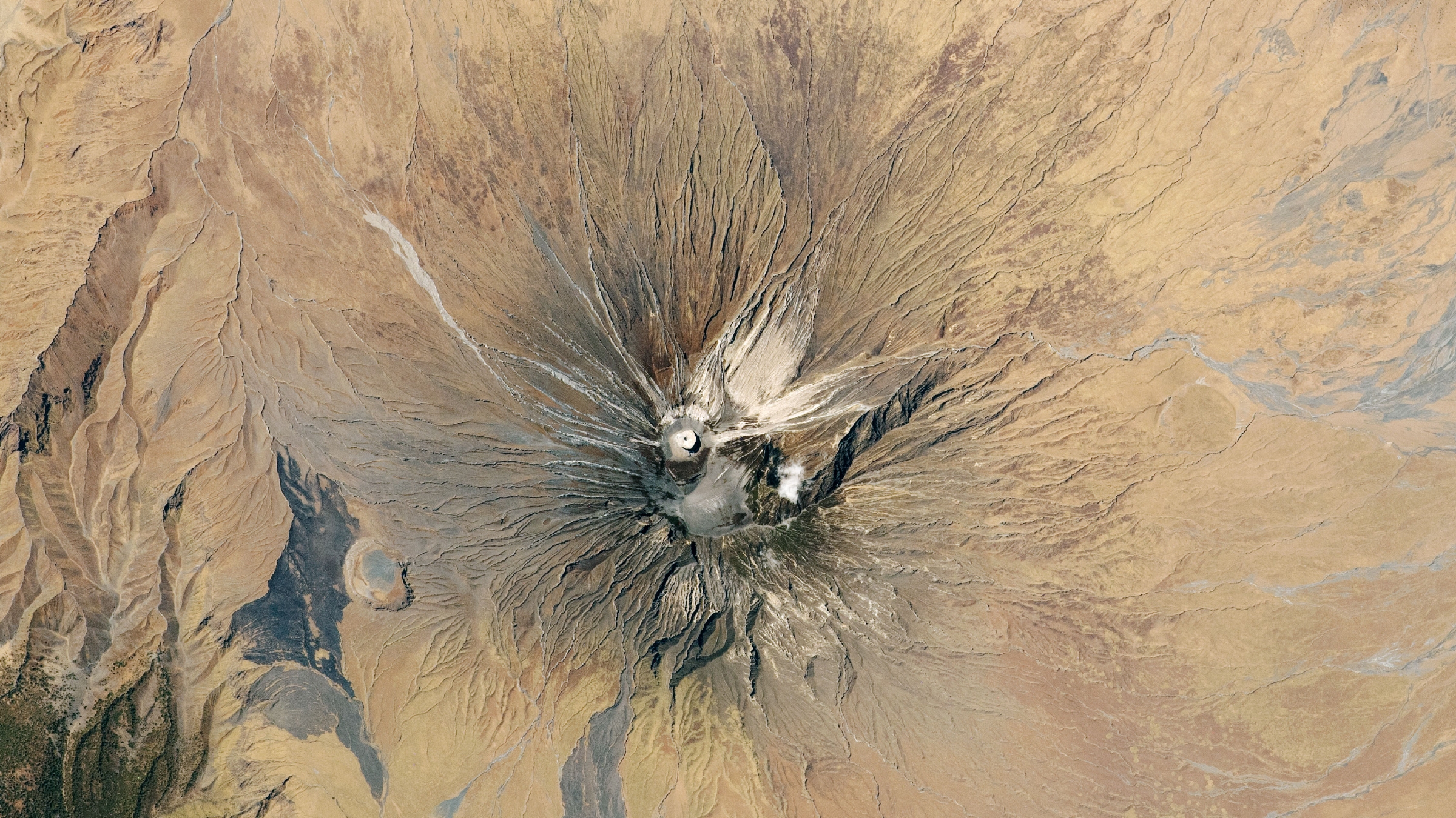 Une photo satellite d'un volcan entouré de plaines arides. Des coulées de lave de couleur claire peuvent être vues sur ses flancs et un cône blanc brillant à son sommet.