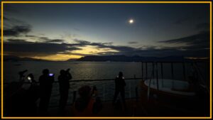 A view of people standing on a beach looking up at a dim sun with a yellow border around the image.